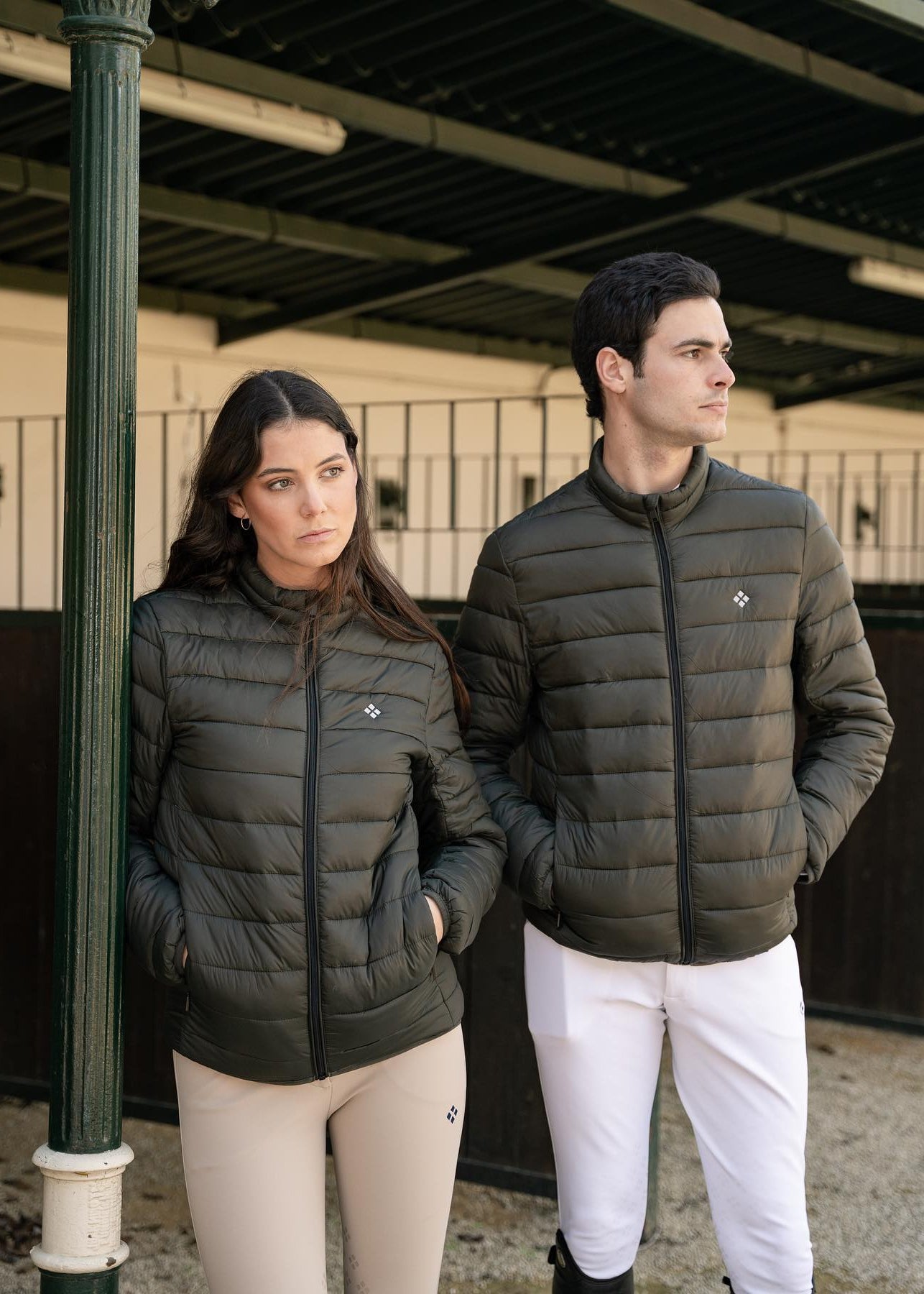 Two people wearing matching green puffer jackets and white pants standing in an equestrian setting.