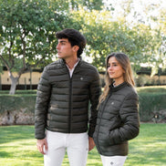 Two people wearing matching dark green puffer jackets and white pants standing in a grassy area with trees.