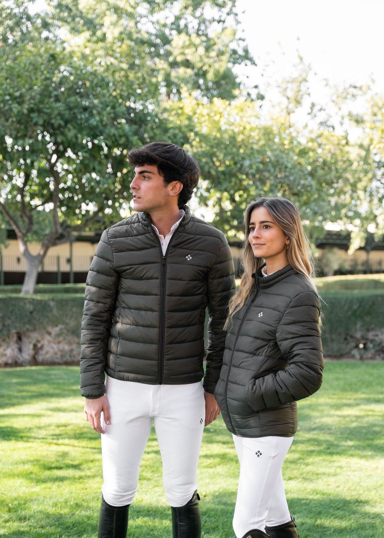 Two people wearing matching dark green puffer jackets and white pants standing in a grassy area with trees.