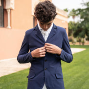 Man wearing a navy blazer over a white shirt outdoors with a building and greenery in the background