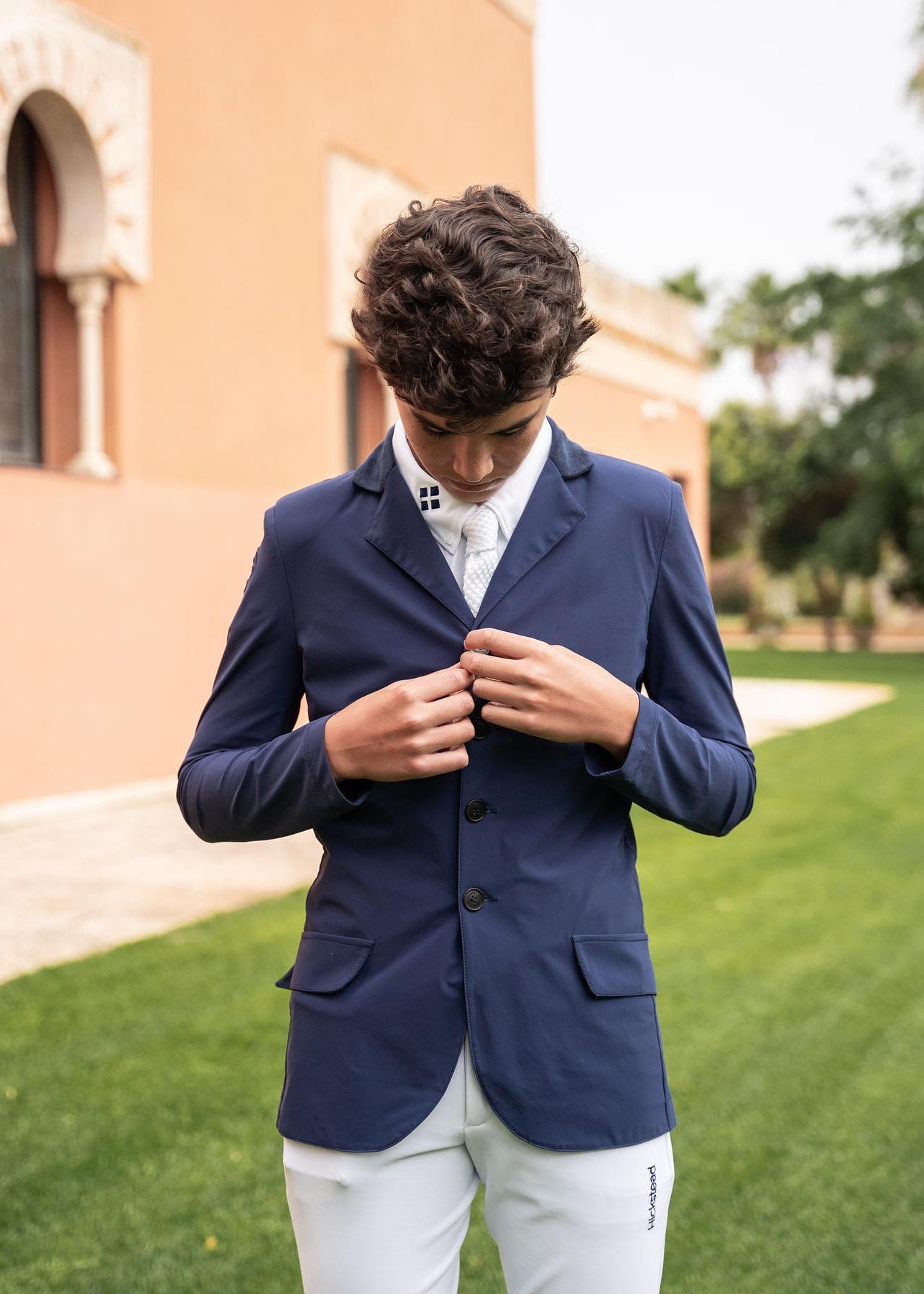 Man wearing a navy blazer over a white shirt outdoors with a building and greenery in the background