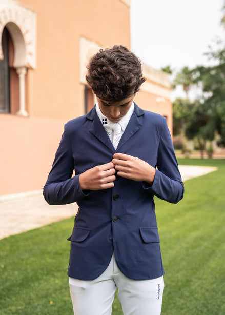 Man wearing a navy blazer over a white shirt outdoors with a building and greenery in the background