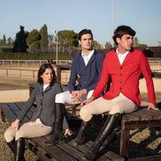 Three people in equestrian attire sitting on a wooden bench at an outdoor setting.