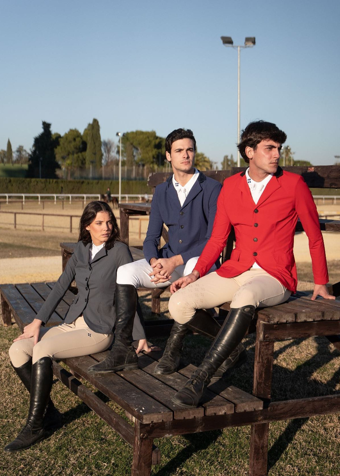 Three people in equestrian attire sitting on a wooden bench at an outdoor setting.