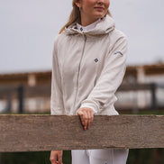 Woman wearing a white equestrian outfit with a logo, standing behind a wooden fence.