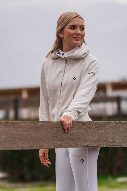 Woman wearing a white equestrian outfit with a logo, standing behind a wooden fence.