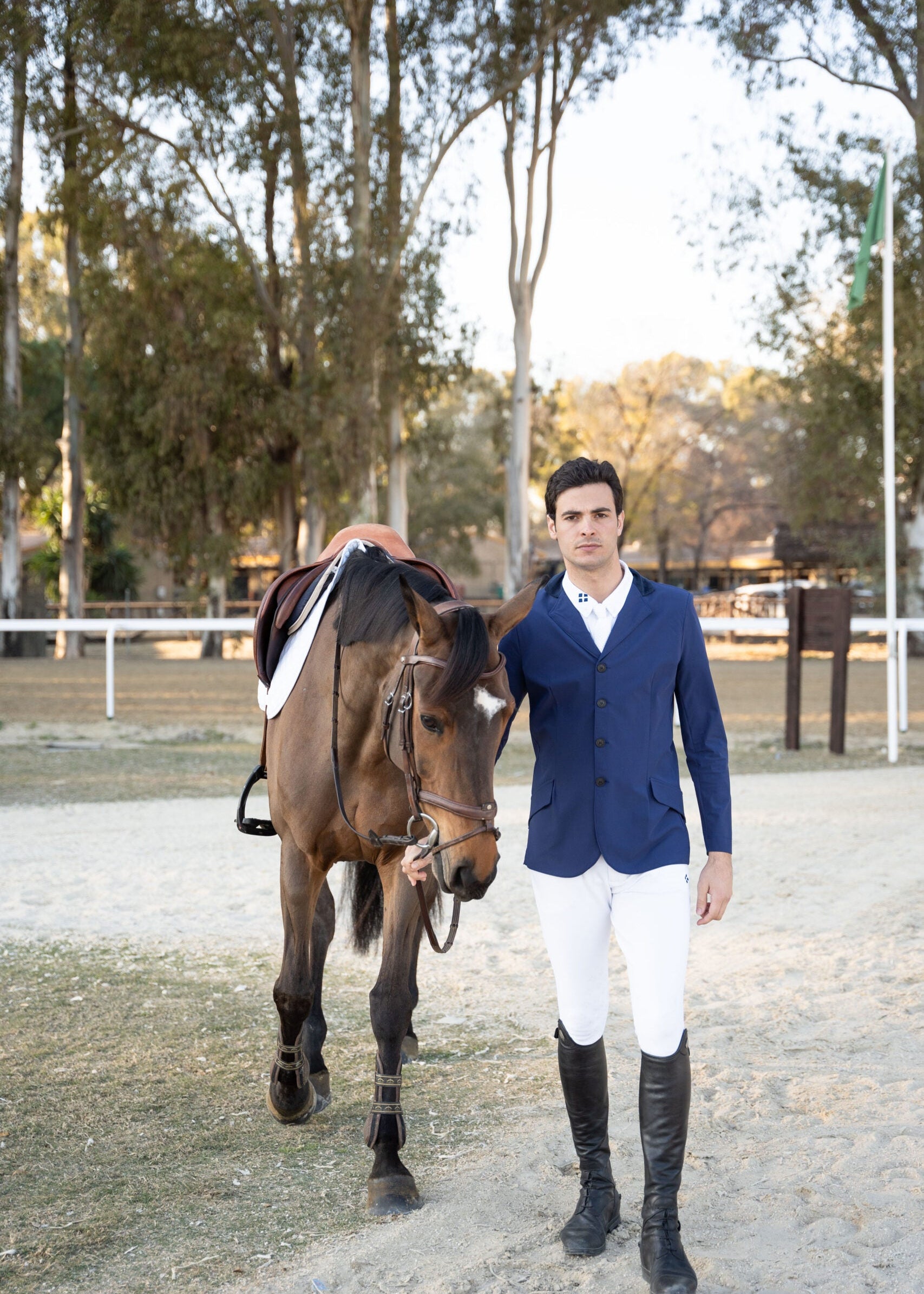 Man in equestrian attire standing next to a horse in an outdoor setting