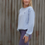 Woman wearing a light gray sweatshirt and purple leggings standing against a wooden wall.