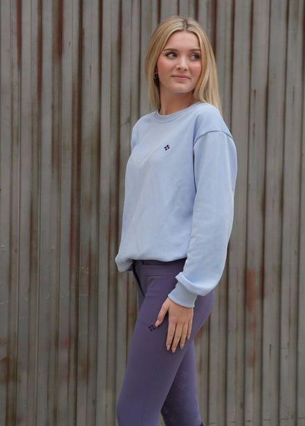 Woman wearing a light gray sweatshirt and purple leggings standing against a wooden wall.